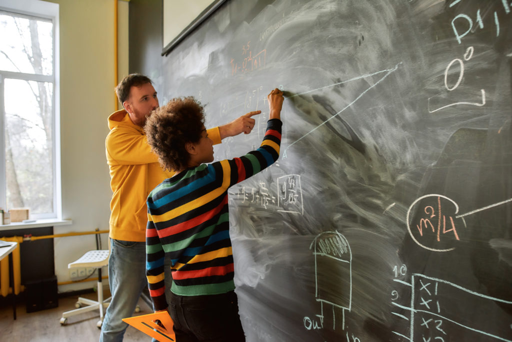 The great aim of education is not knowledge but action. Young male science teacher explaining main rules and formulas in physics and mechanics. A boy is drawing on the blackboard