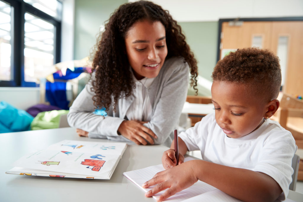 Female infant school teacher working one on one with a young schoolboy, sitting at a table writing in a classroom, front view, close up