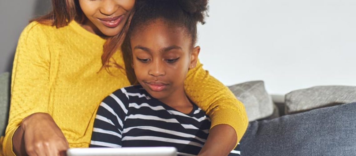 Black mom and daughter learning on tablet sitting at home in sofa