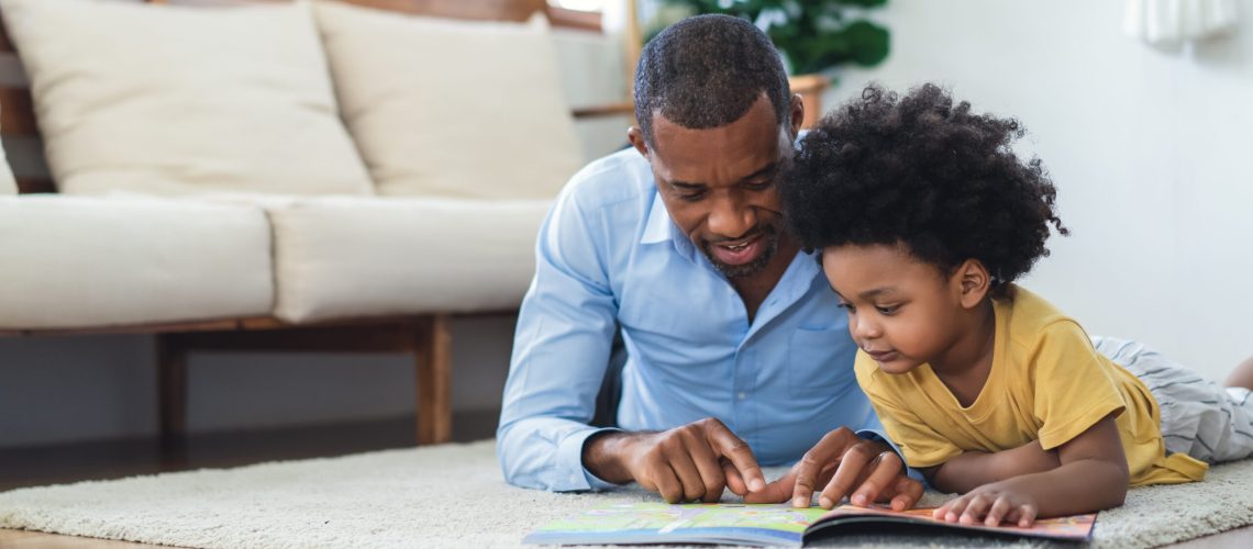 Happy African American father and son are reading a book and smiling while lying on floor spending time together at home. Children education and development concept.