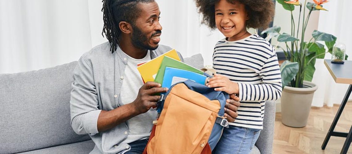 Father's Day. Father helps daughter to put color copybooks in school bag before her first school day. Happy fatherhood