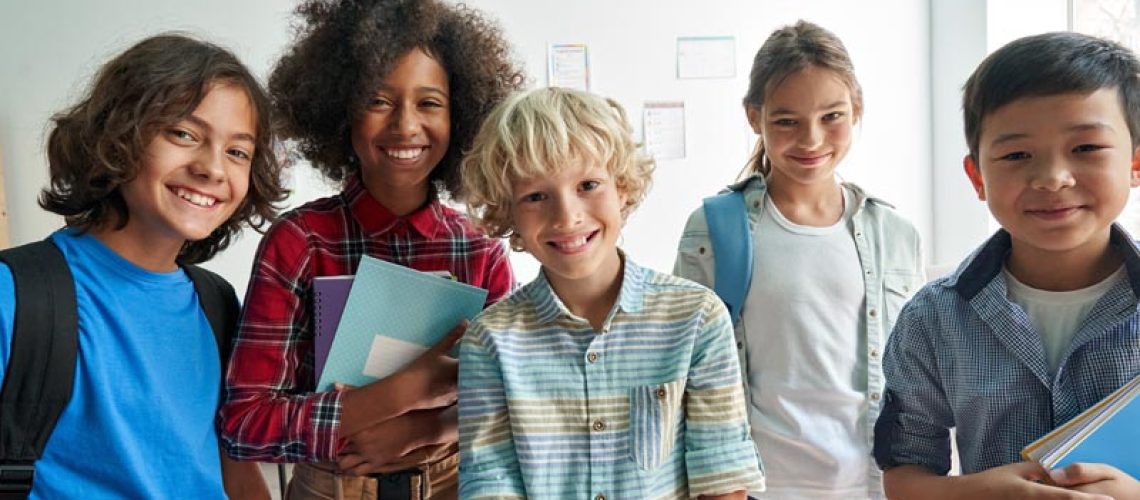 Happy diverse junior school students children group looking at camera standing in classroom. Smiling multiethnic cool kids boys and girls friends posing for group portrait together.