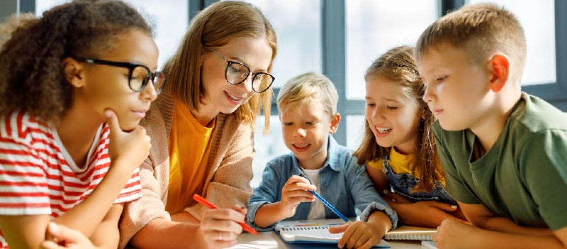 Happy female tutor and optimistic children smiling and discussing topic of   lesson while gathering around table   at school