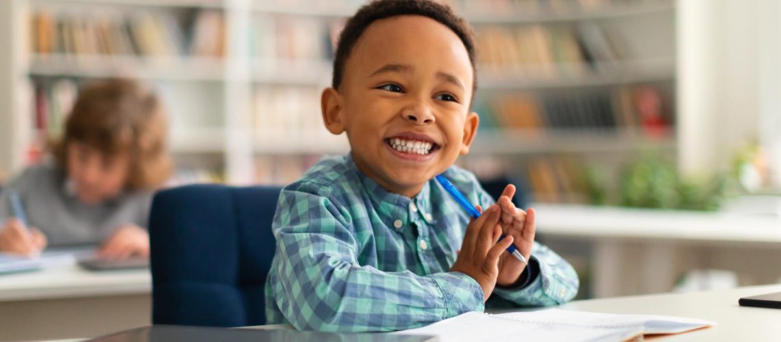 Happy african american primary school boy sitting at desk in classroom, writing in notebook and smiling, free space. Group of diverse classmates studying in the background
