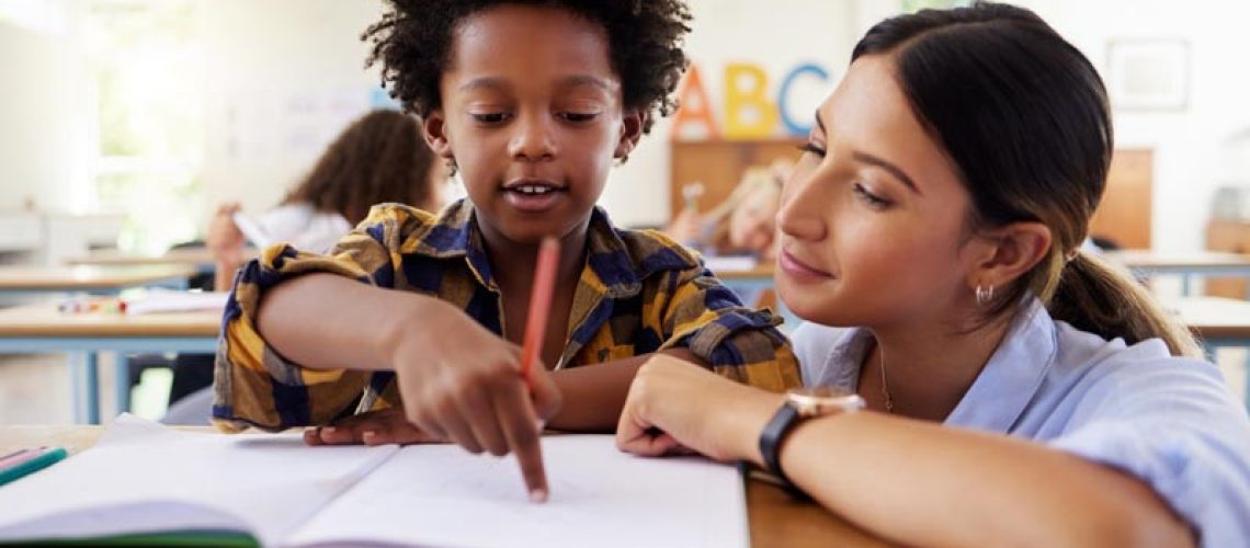 Teacher, learning and helping black kid in classroom for knowledge, studying or assessment. Question, development and boy or student with woman for education pointing in notebook in kindergarten